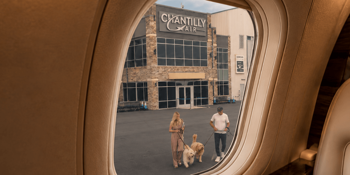 View from inside a private jet cabin overlooking Chantilly Air's FBO terminal, with passengers walking dogs on the ramp outside.