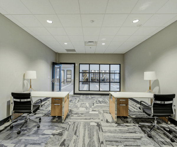 Furnished private office with dual desks, ergonomic chairs, large interior windows, and natural light inside a full-service jet center office space.