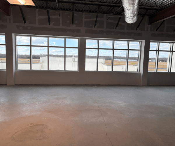 Third-floor open office shell with concrete floors and large windows overlooking an active airport runway and aviation facilities.